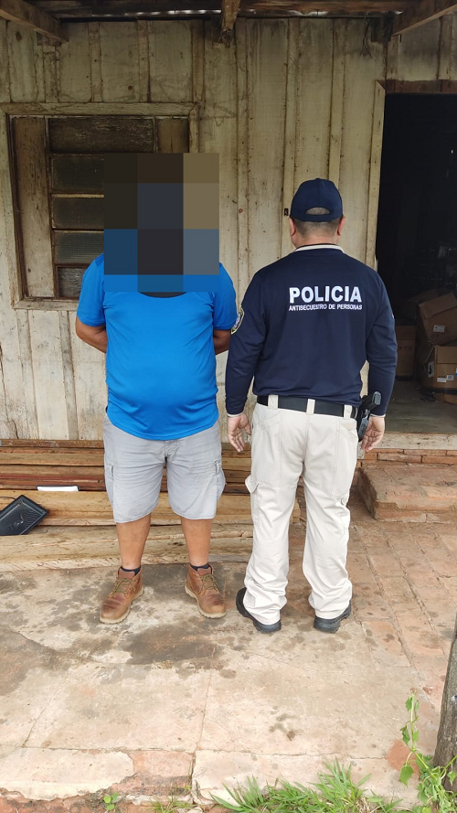 A man whose face has been disguised stands handcuffed next to a Paraguayan law enforcement officer.